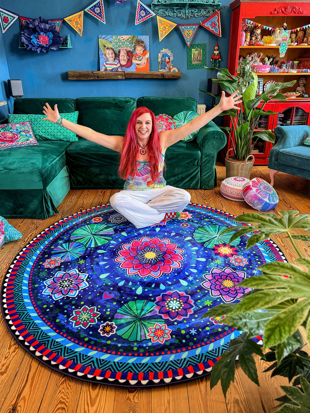 Bine Brändle sitzt lächelnd im Schneidersitz auf einem runden Teppich mit floralem Mandala-Muster in Dunkelblau, Pink, Türkis, Grün und Rot und breitet die Arme weit aus. Der Teppich liegt auf warmem Holzboden in einem gemütlichen Wohnzimmer mit grünem Samtsofa, Zimmerpflanzen, bunten Kissen und dekorativen Möbeln vor einer dunkelblauen Wand. Die Szene wirkt fröhlich, kreativ und einladend mit lebendigem Boho-Wohnstil.