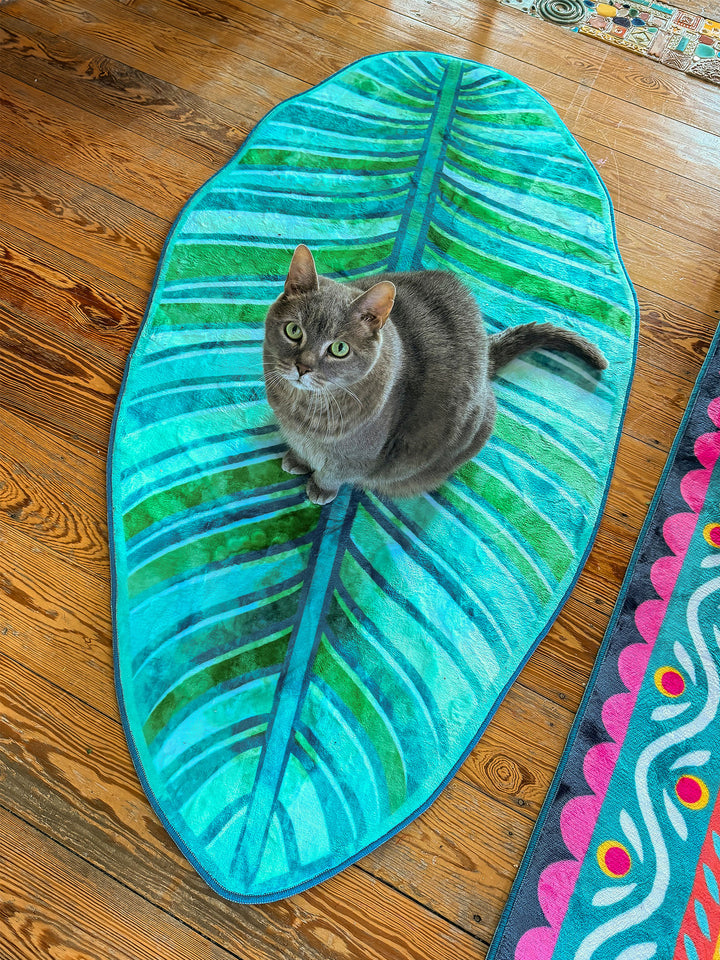 Graue Katze sitzt aufmerksam auf einem grünen Blatt-Teppich mit tropischem Blattdesign in Türkis, Blau und verschiedenen Grüntönen. Der Teppich liegt auf warmem Holzboden neben einem weiteren bunten Teppich mit floralen Mustern. Die Katze blickt direkt in die Kamera und verleiht der Szene eine gemütliche, wohnliche Atmosphäre mit kreativem und naturinspiriertem Stil.