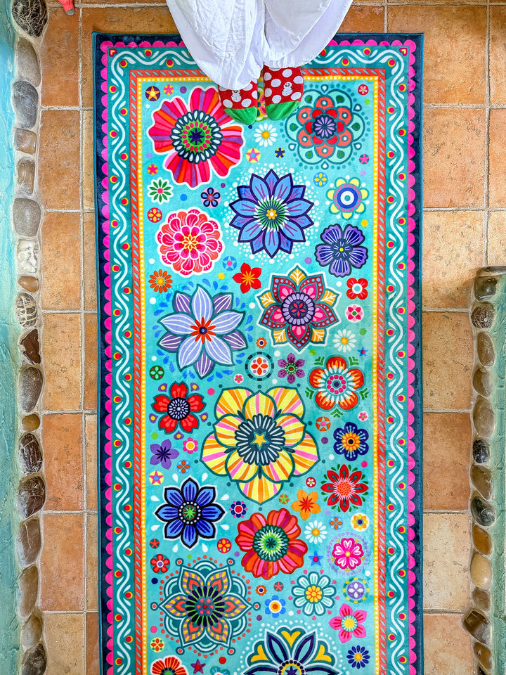 Schmaler rechteckiger Badteppich in Türkis mit farbenfrohen Blumen- und Mandalamotiven in Pink, Blau, Gelb, Rot und Grün liegt auf terrakottafarbenen Fliesen vor einer Badewanne. Das kreative Badezimmer ist mit kunstvollen Mosaikwänden, Naturstein-Details und Pflanzen dekoriert. Große grüne Blätter und ein Fenster mit Tageslicht schaffen eine frische, natürliche Atmosphäre. Der lebendige Teppich setzt einen fröhlichen Farbakzent im individuell gestalteten Boho-Bad.