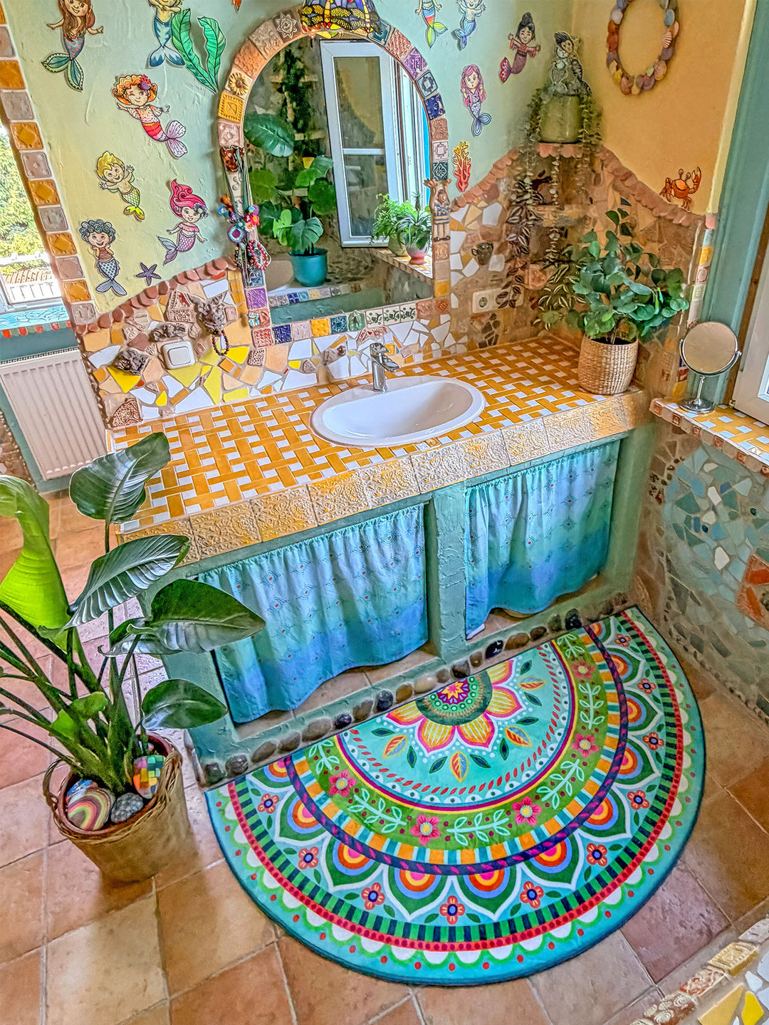 Halbrunder Badteppich in frischen Türkis-, Grün-, Pink- und Gelbtönen liegt auf beigefarbenen Fliesen vor einem kunstvoll gestalteten Waschbecken im Boho-Stil. Das Teppichdesign zeigt ein großes Mandala mit floralen Ornamenten, Blütenranken und bunten Bordüren. Darüber befindet sich ein Waschtisch mit gelb-weißer Mosaikplatte, integriertem Waschbecken und türkisfarbenen Stoffvorhängen als Unterschrank. Die Wände sind mit Naturstein, bunten Fliesen, Spiegelbogen und verspielten Meerjungfrauen-Motiven dekorie