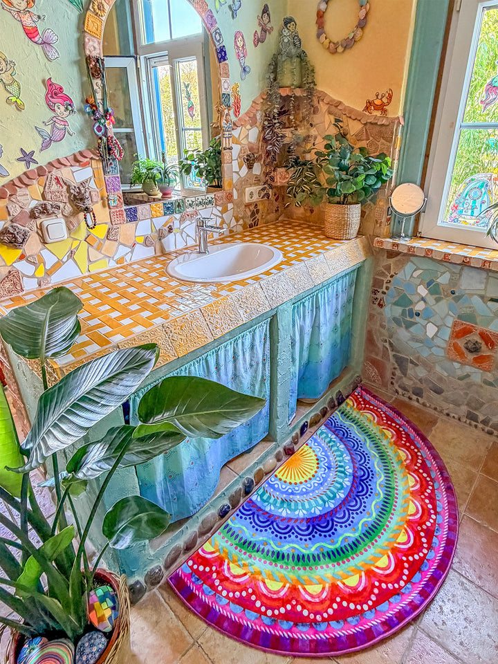 Halbrunder Teppich mit farbenfrohem Mandala-Muster in Blau, Türkis, Gelb, Rot, Pink und Lila liegt vor einem Waschbecken in einem kunstvoll gestalteten Badezimmer. Der Raum ist mit Mosaikfliesen, Spiegelbogen, Pflanzen und verspielten Wanddekorationen geschmückt. Große grüne Blätterpflanzen und warme Naturtöne schaffen eine kreative, mediterrane Wohlfühlatmosphäre. Der bunte Badteppich setzt einen lebendigen Akzent im individuellen Badezimmerdesign.
