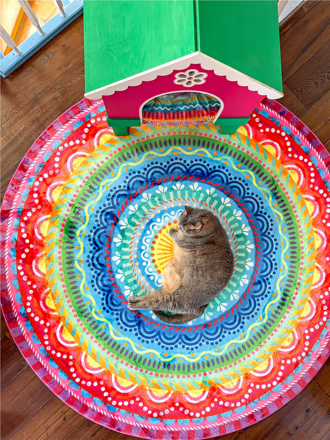 Graue Katze liegt entspannt auf einem runden Mandala-Teppich mit leuchtenden Farben in Blau, Türkis, Grün, Gelb, Orange, Pink und Rot. Der bunte Teppich liegt auf dunklem Holzboden neben einem verspielten Katzenhaus in Pink und Grün. Kreisförmige Muster und kräftige Farben schaffen eine fröhliche, kreative Wohnatmosphäre mit gemütlichem Platz für Haustiere.