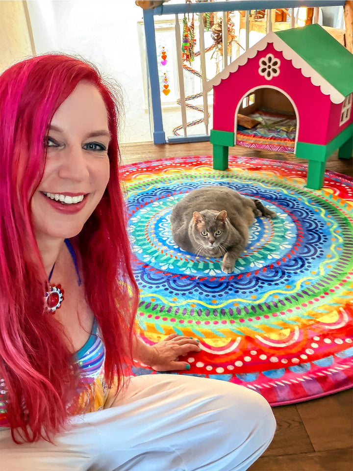 Bine Brändle sitzt lächelnd neben einer grauen Katze auf einem runden, farbenfrohen Mandala-Teppich in Blau, Türkis, Rot, Gelb und Pink. Die Katze liegt entspannt in der Mitte des Teppichs und blickt in die Kamera. Im Hintergrund steht ein buntes Katzenhaus in Pink und Grün auf Holzfußboden. Die fröhliche Wohnszene verbindet kreative Farben, gemütliches Wohnen und verspieltes Design.